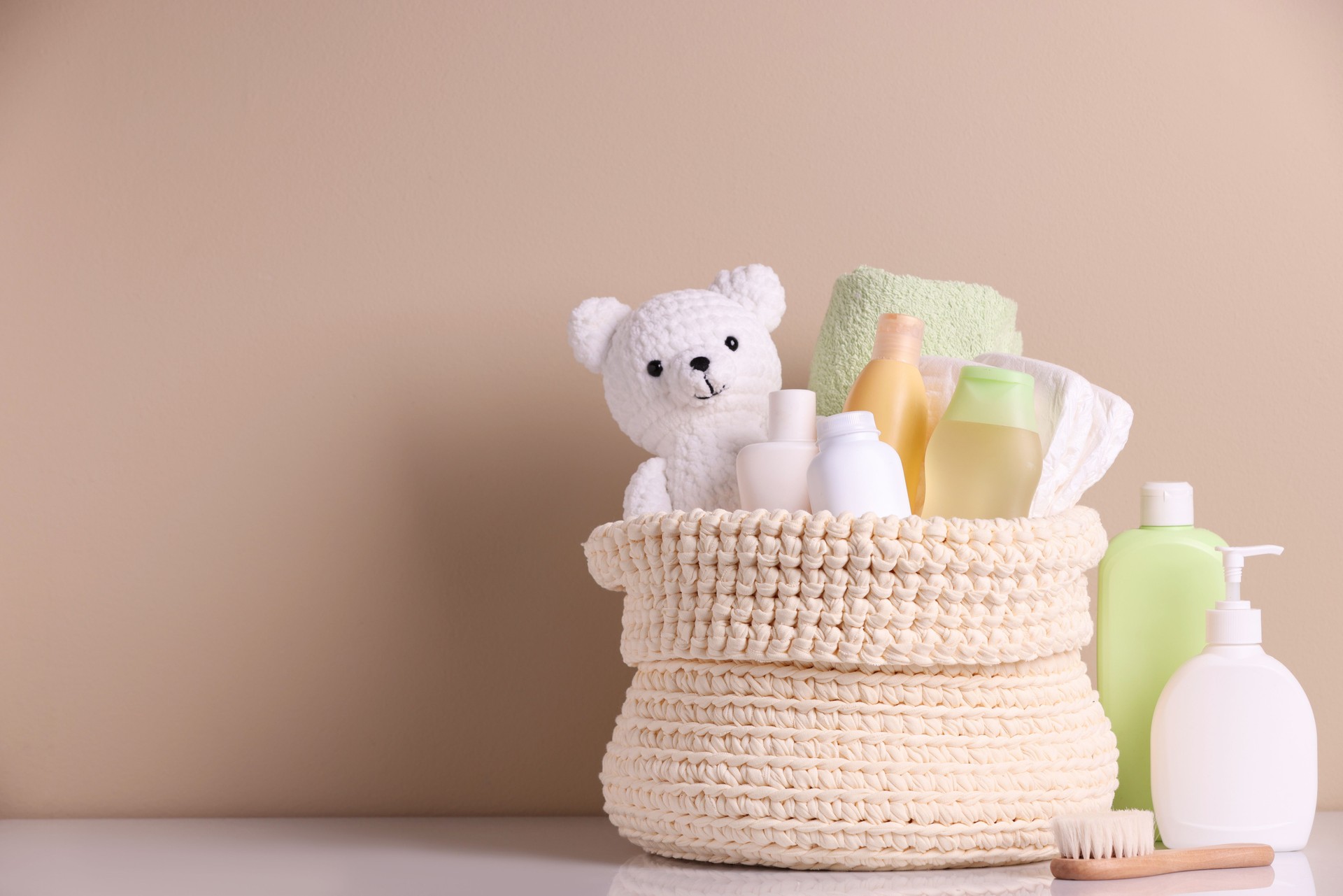 Knitted basket with baby cosmetic products, bath accessories and toy bear on white table against beige background. Space for text